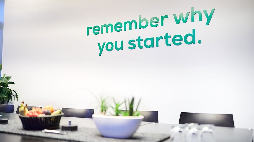 Das Foto zeigt ein Meetingraum mit verschiedenen Tischdekorationen. Auf der Wand Im Hintergrund steht "remember why you started.".